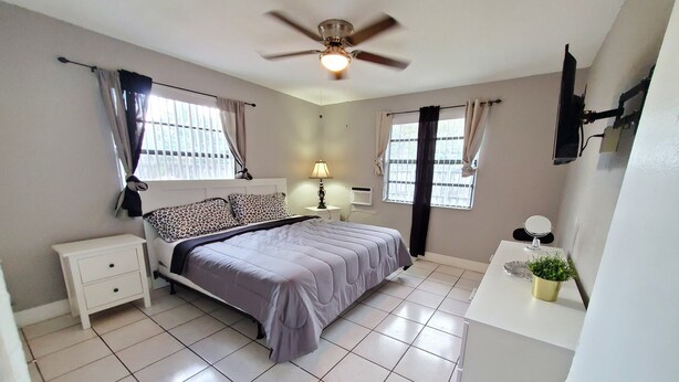 A bedroom with a bed, nightstand, lamp, window, ceiling fan, and a small potted plant. - Furnished 2BR - Ideal for Relocation & Extended Stays (Fort Lauderdale)
