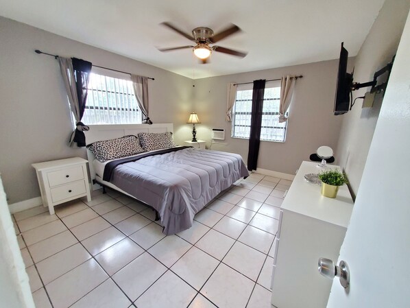 A bedroom with a bed, nightstand, lamp, window, ceiling fan, and a small potted plant. - Furnished 2BR - Ideal for Relocation & Extended Stays (Fort Lauderdale)