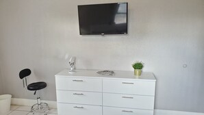A bathroom with a white vanity, a mirror, a small potted plant, a black chair, and a flat-screen TV mounted on the wall. - Furnished 2BR - Ideal for Relocation & Extended Stays (Fort Lauderdale)