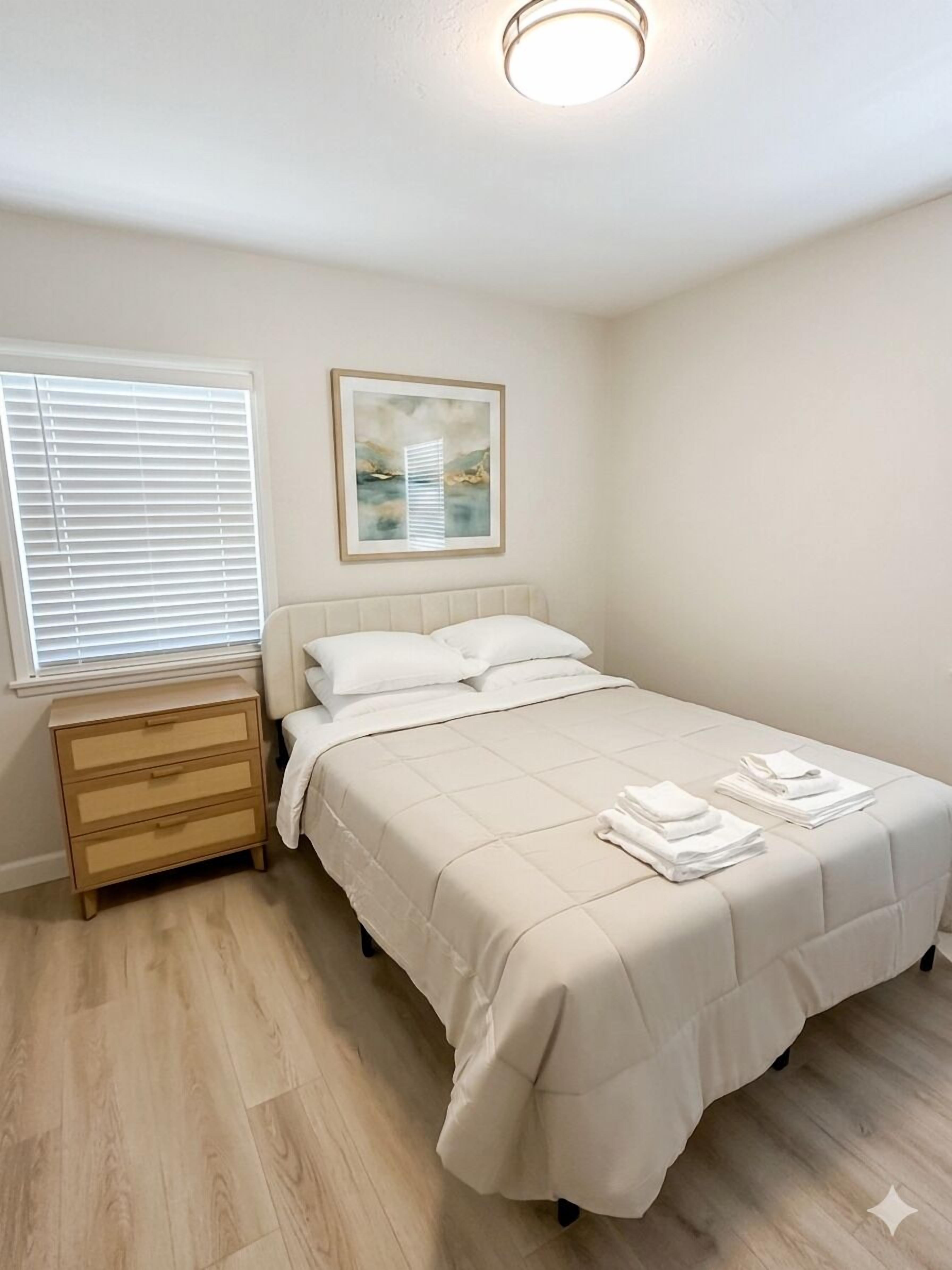 Une chambre à coucher avec un lit, une table de chevet, une fenêtre avec des stores et un tableau encadré au mur.