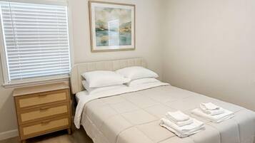 Une chambre à coucher avec un lit, une table de chevet, une fenêtre avec des stores et un tableau encadré au mur.