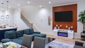 A modern living room with a sectional sofa, a flat-screen TV mounted on a wooden wall, a fireplace, and a staircase leading to another level. - 10 Mins to NRG, Sleeps 12, Hot Tub, 4BR Home (Houston)