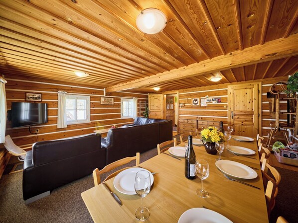 A wooden cabin interior with a dining table set for two, a sofa, and a television mounted on the wall.