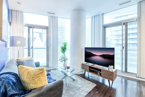 A modern living room with a glass coffee table, a sofa with cushions, a flat-screen TV, and large windows with a city view.