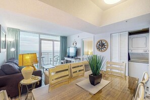 A room with a dining table, chairs, a television, a clock, and a window with curtains. - Oceanfront 1BR Condo | Balcony, Pool & Views (Myrtle Beach)