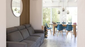 En moderne stue med en grå sofa, et spiseområde med et træbord og blå stole, samt et stort vindue med naturligt lys.