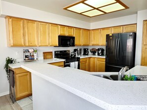 A modern kitchen with wooden cabinets, a black refrigerator, a microwave, a stove, and a coffee maker. - Fully Furnished 3BR Home in Gated East Mesa (Mesa)