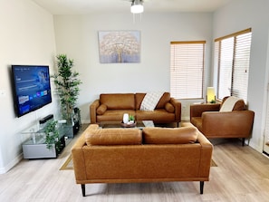A living room with a brown sofa, a glass coffee table, a TV mounted on the wall, and a painting of a tree. - Fully Furnished 3BR Home in Gated East Mesa (Mesa)