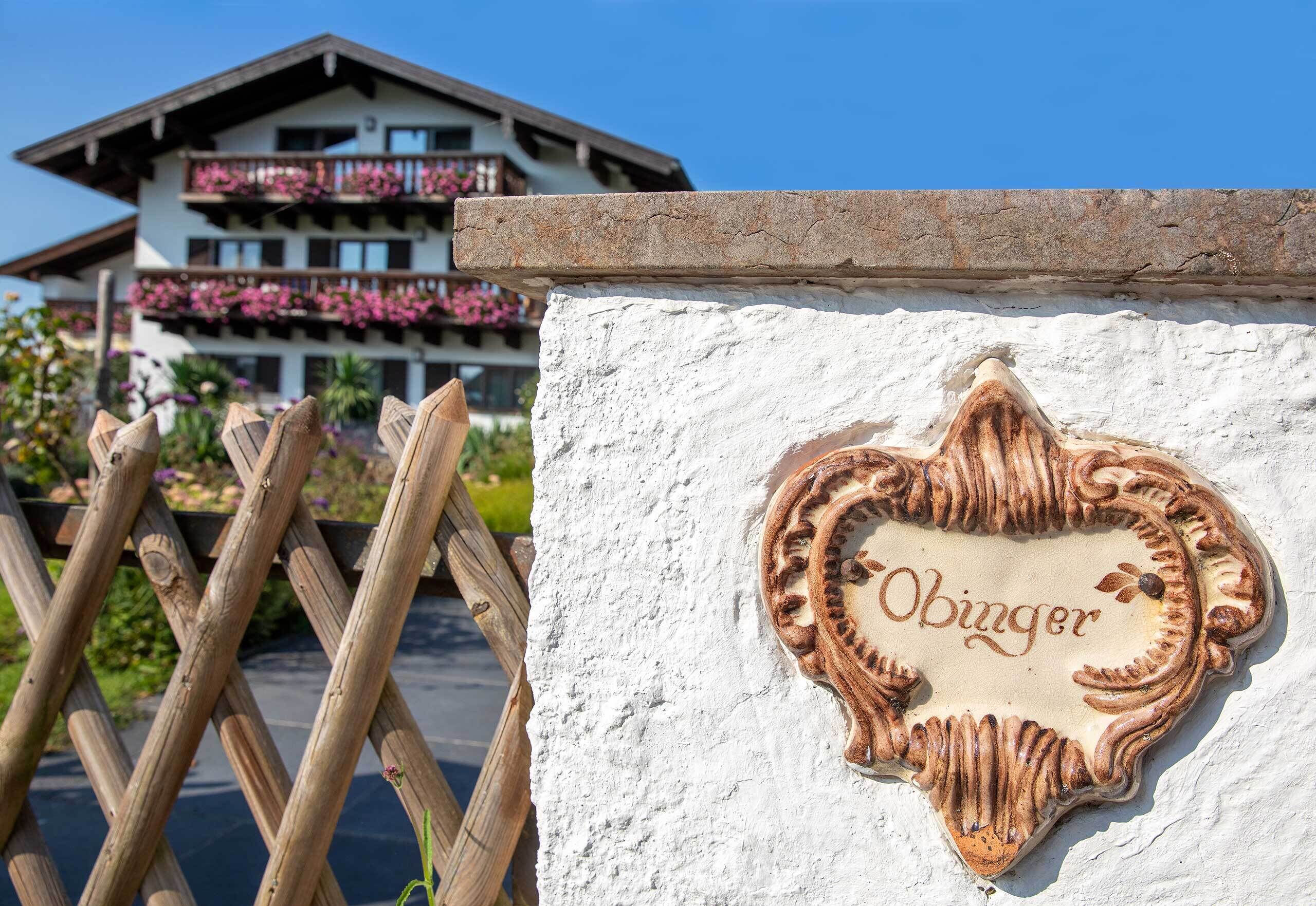 A heart-shaped sign with 'Obinger' on a white wall, a wooden fence, and a building with balconies in the background.