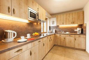 A modern kitchen with wooden cabinets, stainless steel appliances, and a countertop with a kettle, plates, and a fruit bowl. - New four star apartment no. 7 with 52 square meters, Die Bequeme (Gstadt am Chiemsee)