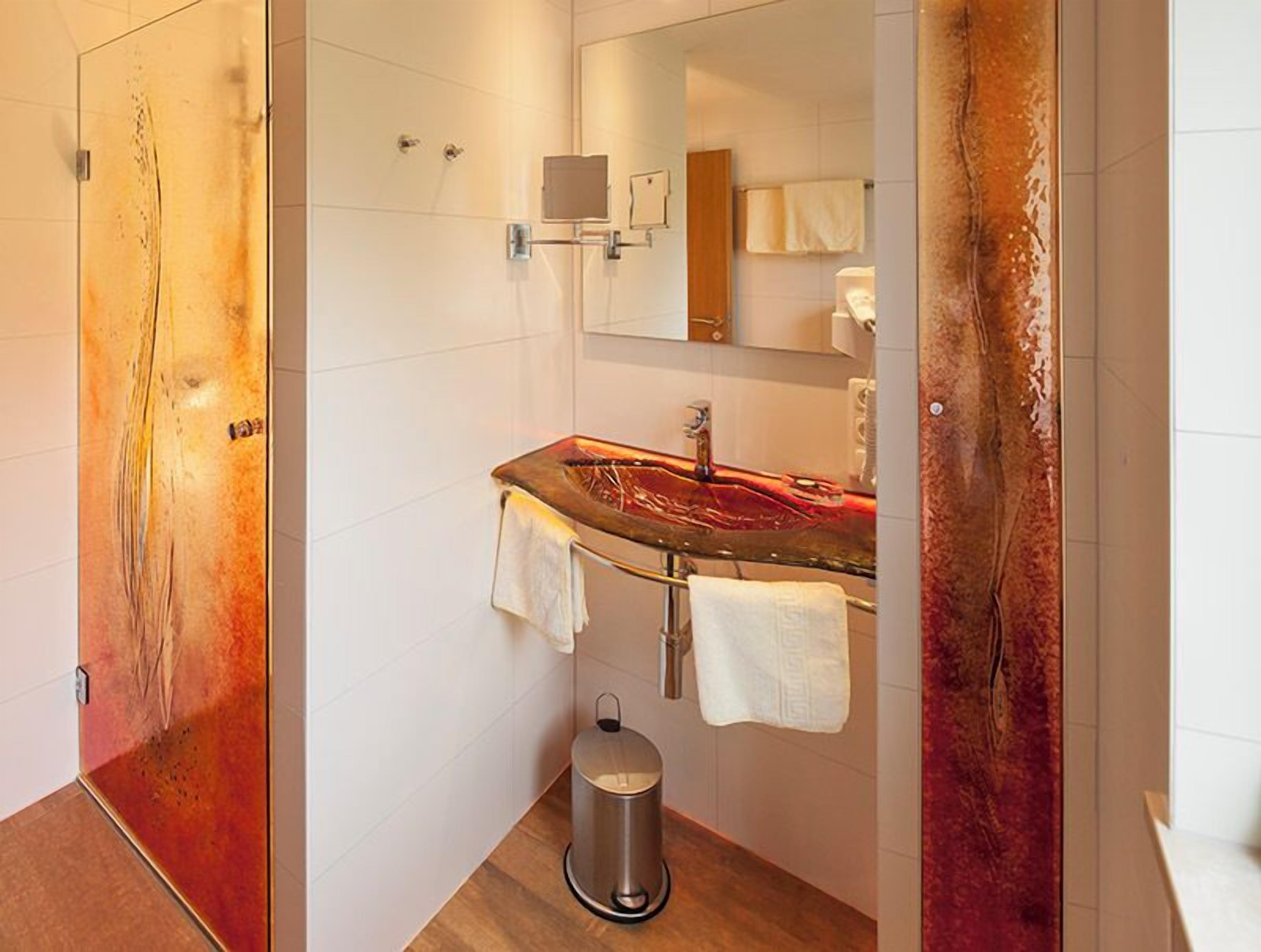 A modern bathroom with a glass shower enclosure, a stainless steel sink, and a trash bin.