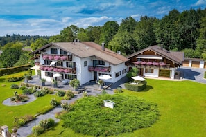 A two-story chalet with a balcony, surrounded by a well-maintained garden and a forested backdrop. - New four star apartment no. 7 with 52 square meters, Die Bequeme (Gstadt am Chiemsee)