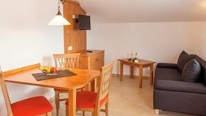 A cozy living room with a dark sofa, a dining table with chairs, a small side table, and a TV mounted on a wooden wall. - New four-star vacation apartment with 42 sqm No. 3, The Sunny One (Gstadt am Chiemsee)