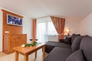 Ein Wohnzimmer mit einer Couch, einem Holzkasten, einem Fernseher, einem Tisch mit Weingläsern und einem Fenster mit Vorhängen.