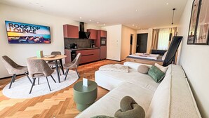 Ein modernes Wohnzimmer mit einer Couch, Sesseln, einem Couchtisch und einem Fernseher. Es gibt eine Küchenzeile mit Schränken und einem Esstisch.
