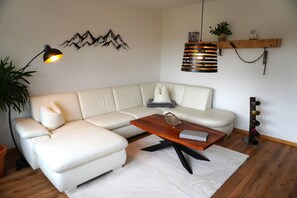 A modern living room with a white leather sofa, a wooden coffee table, a black floor lamp, a wall-mounted lamp, and a wine rack. - Vacation apartment "Wendelsteinblick" (Heufeld)