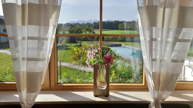 A window with a view of a swimming pool, greenery, and distant mountains. - Vacation apartment "Wendelsteinblick" (Heufeld)