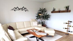 A living room with a sectional sofa, a wooden coffee table, a potted plant, a wall-mounted wine rack, and a decorative wall piece. - Vacation apartment "Wendelsteinblick" (Heufeld)