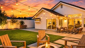 Un patio trasero con un brasero, mobiliario exterior e iluminación colgante bajo un cielo de atardecer.