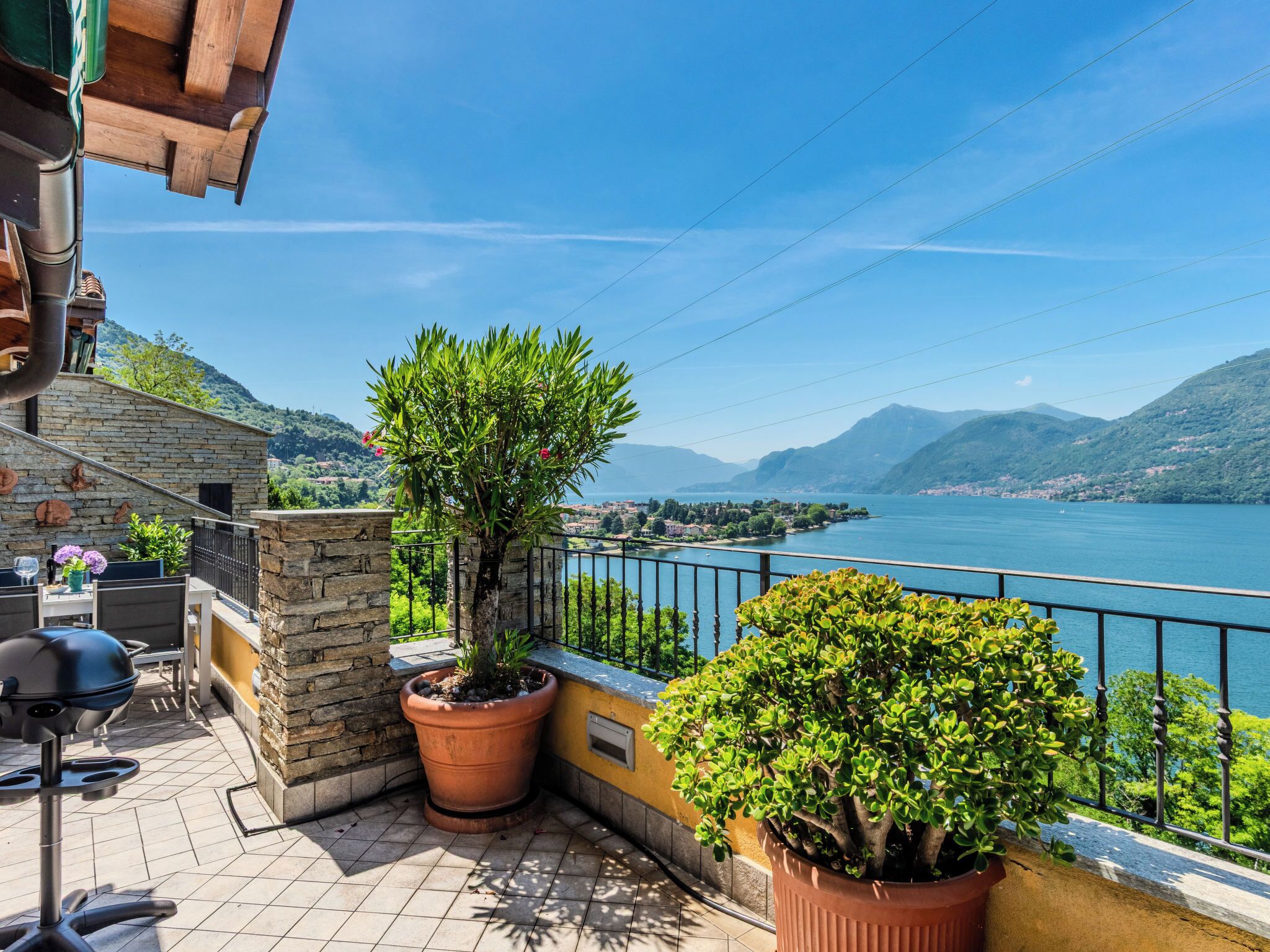 A terrace with a stone wall, potted plants, a grill, and a view of a lake and mountains.