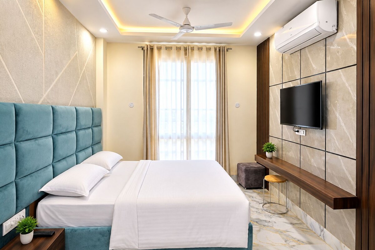 A modern hotel room with a large bed, a flat-screen TV mounted on the wall, a ceiling fan, and a large window with curtains.