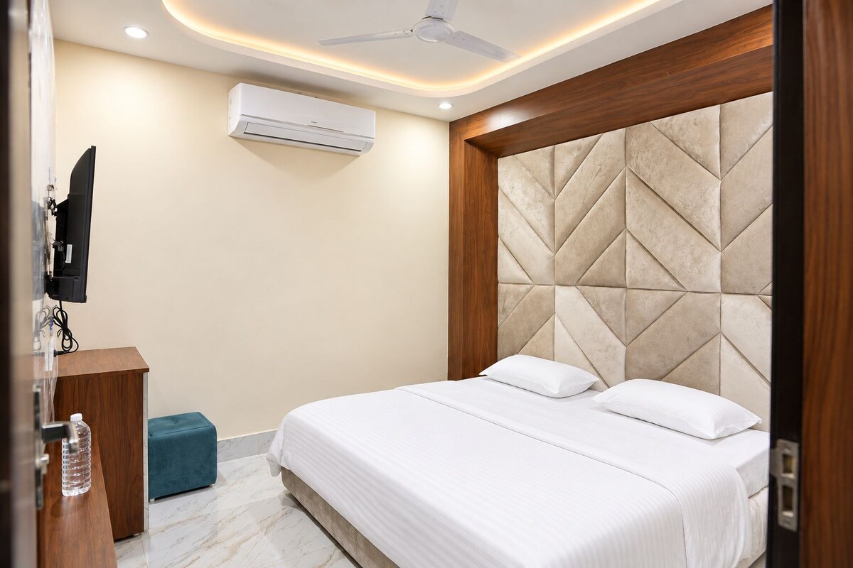A hotel room with a bed, a wooden headboard, an air conditioning unit, a television, and a small blue ottoman.