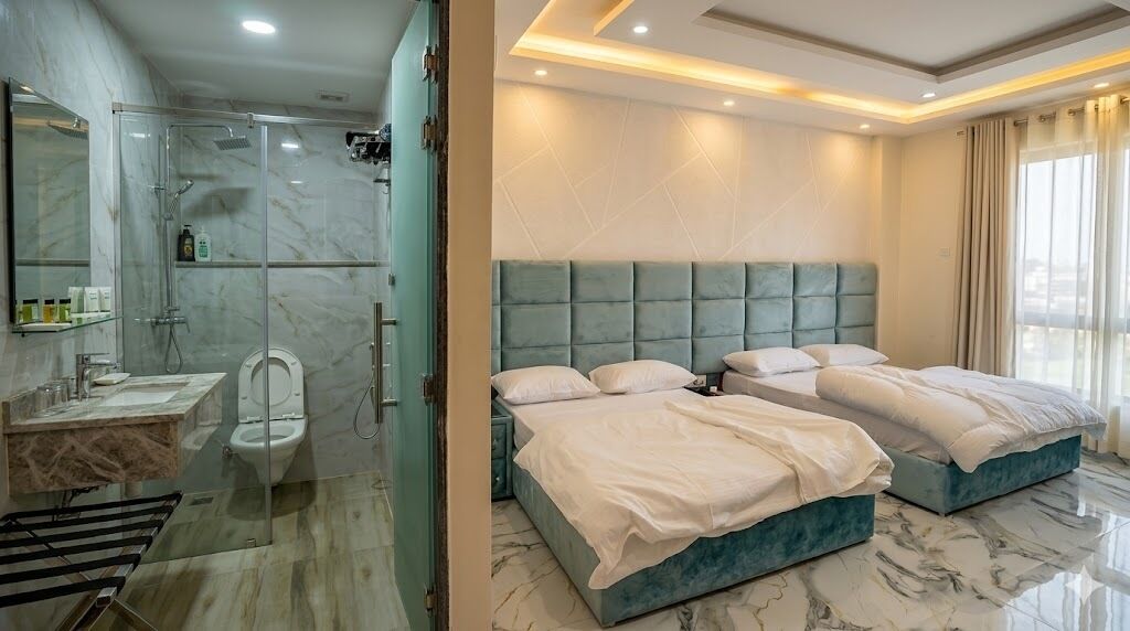 A modern bathroom with a glass shower enclosure, a marble-tiled floor, and a separate toilet. The bedroom features two beds with a teal headboard, a large window, and a marble floor.