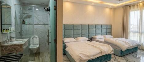 A modern bathroom with a glass shower enclosure, a marble-tiled floor, and a separate toilet. The bedroom features two beds with a teal headboard, a large window, and a marble floor.