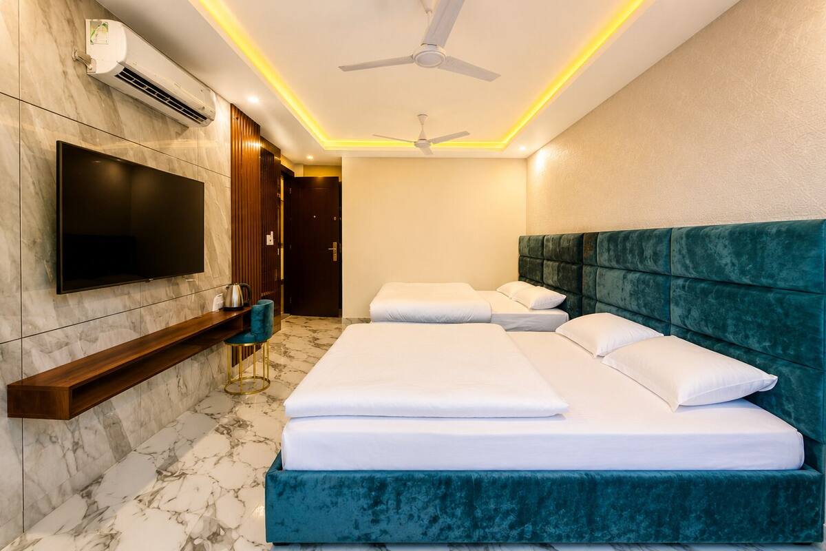 Habitación de hotel con televisión, ventilador de techo, estante de madera, piso de mármol y cabecera tapizada en verde.