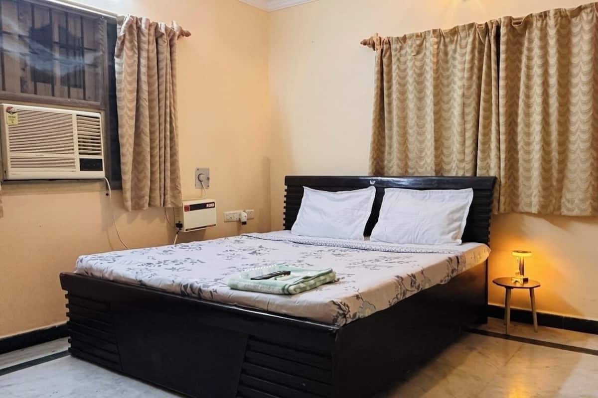 A bedroom with a bed, curtains, an air conditioner, a bedside lamp, and a wall socket.