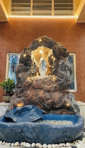 Une formation rocheuse abritant une statue dans une niche, le tout entouré d’un élément décoratif aquatique et de plantes en pot.