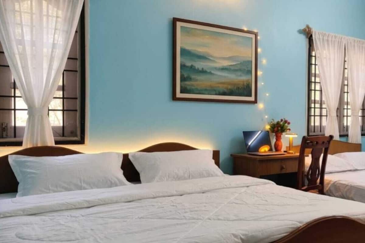 A room with two beds, a desk with a lamp, a painting on the wall, and a window with curtains.