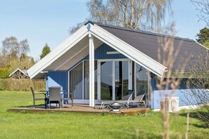 Et hus med skråt tag, store glasdøre, en terrasse med stole og en klimaanlæg.