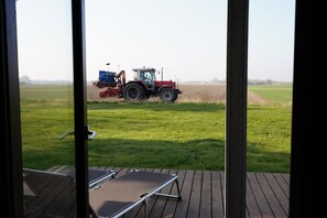Une vue à travers une fenêtre donnant sur une terrasse avec une chaise, un tracteur dans un champ et des éoliennes au loin.