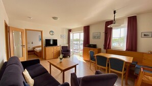 A living room with a sofa, a TV, a wooden cabinet, and a dining area with chairs and a table. - Ferienwohnung in Orth auf Fehmarn (Fehmarn)