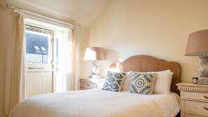 Ein Bett mit weißer Bettdecke, zwei Zierkissen, ein hölzernes Kopfende, ein Nachttisch mit Lampe und ein Fenster mit Vorhängen.