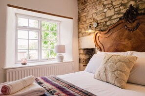 A bedroom with a large window, a stone wall, a wooden headboard, a bedside lamp, and a rolled towel on the bed. - Beautiful 3 Bedroom Cottage in the Cotswolds (Milton-under-Wychwood)