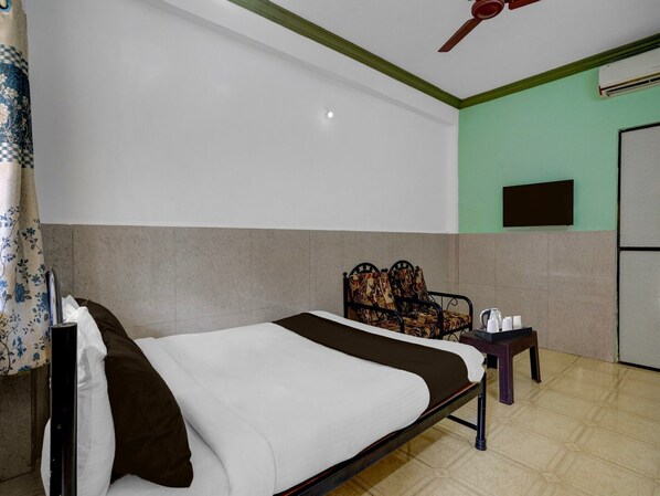A hotel room with a bed, a chair, a table with a kettle, a television, and a ceiling fan.