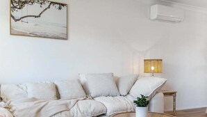 Un salon comprenant un canapé, une table basse ronde en bois, une petite table d’appoint, un climatiseur fixé au mur et un tableau encadré accroché au mur.