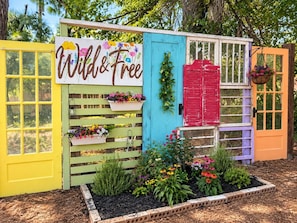Una estructura colorida al aire libre con un letrero que dice 'Wild & Free', que presenta una valla de madera, macetas con flores y una pequeña área de jardín.