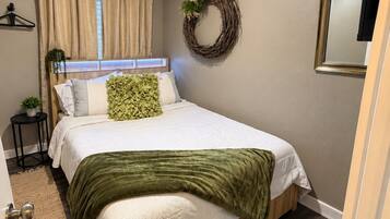 A bedroom with a bed, a ceiling fan, a window with curtains, a wall-mounted mirror, and a door.