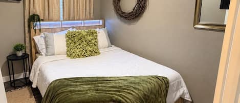 A bedroom with a bed, a ceiling fan, a window with curtains, a wall-mounted mirror, and a door.