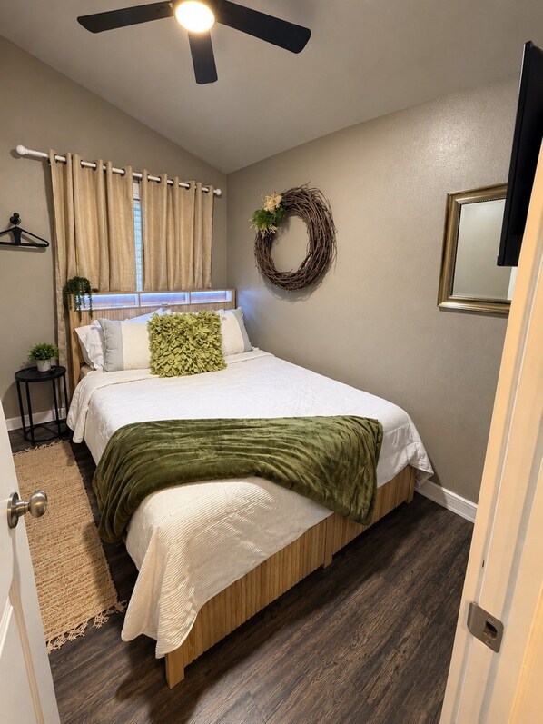 A bedroom with a bed, a ceiling fan, a window with curtains, a wall-mounted mirror, and a door.