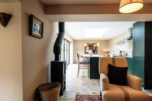 A cozy living room with a fireplace, a sofa, and a kitchen in the background. - The Potting Shed Banham Cottages (Bourton-on-the-Water)