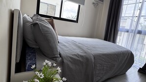 Un dormitorio con cama, mesita de noche con planta, ventana con cortinas y aire acondicionado de pared.
