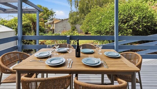 A wooden table set for four with wicker chairs, a bottle of wine, and a view of greenery and a house in the background. - Seednest Blue Lake King Bed & BBQ Backyard (Mount Gambier)