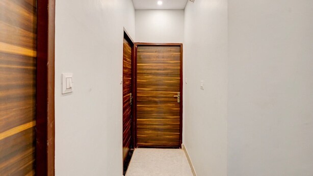 Un pasillo estrecho con una puerta de madera al final, paredes blancas y un interruptor de luz a la izquierda.