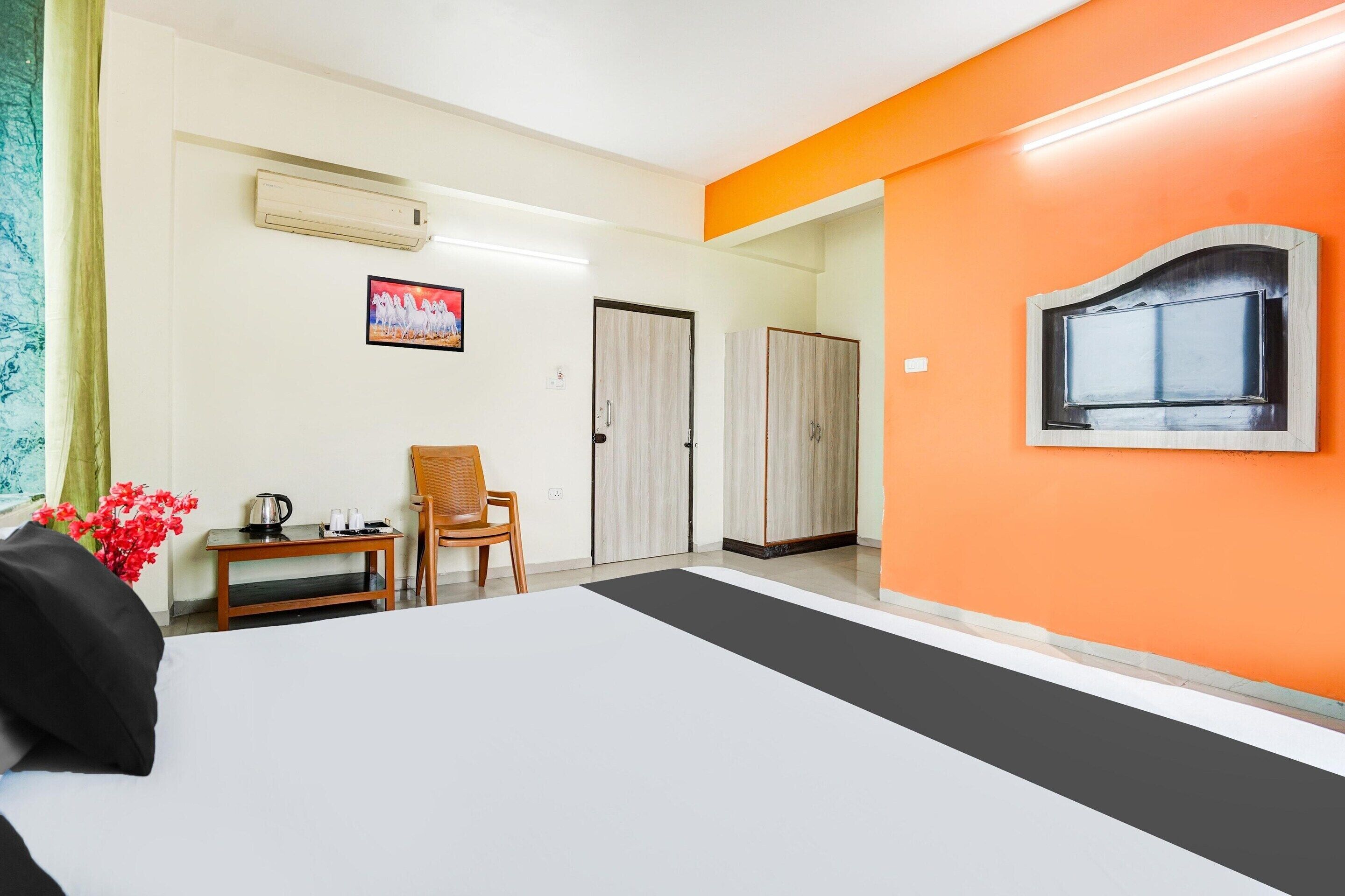 Une chambre d’hôtel comprenant un lit, un bureau avec une chaise, un appareil de climatisation, un tableau encadré et un mur d’accent de couleur orange.
