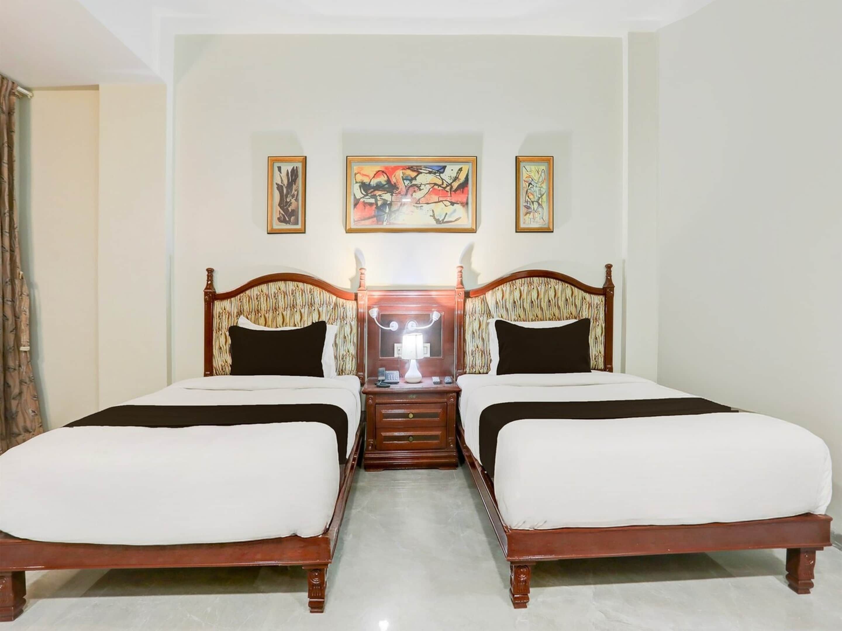 A double bed with a wooden frame and a headboard with a patterned design. There is a nightstand with a lamp and a mirror. The room has plain walls and a light-colored floor.
