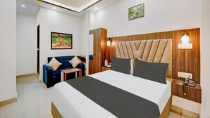 A modern hotel room with a large bed, a wooden headboard, a nightstand with a phone, a blue sofa, and a painting on the wall. - Premium Anisabad Near Mahavir Sansthan (Patna)